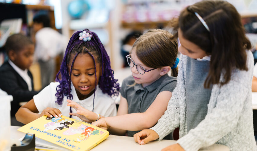 Three Elementary school students sit at a table and read a French children's book