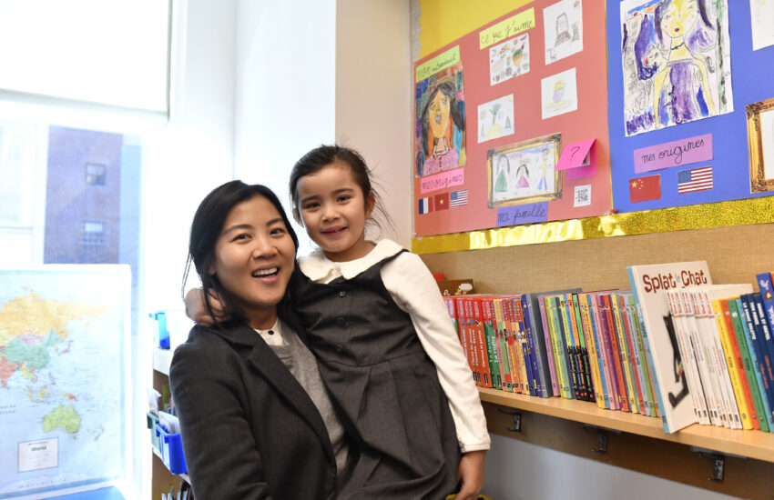 Mother and daughter in the classroom smile in front of a poster that describes the child's family and countries of origin.